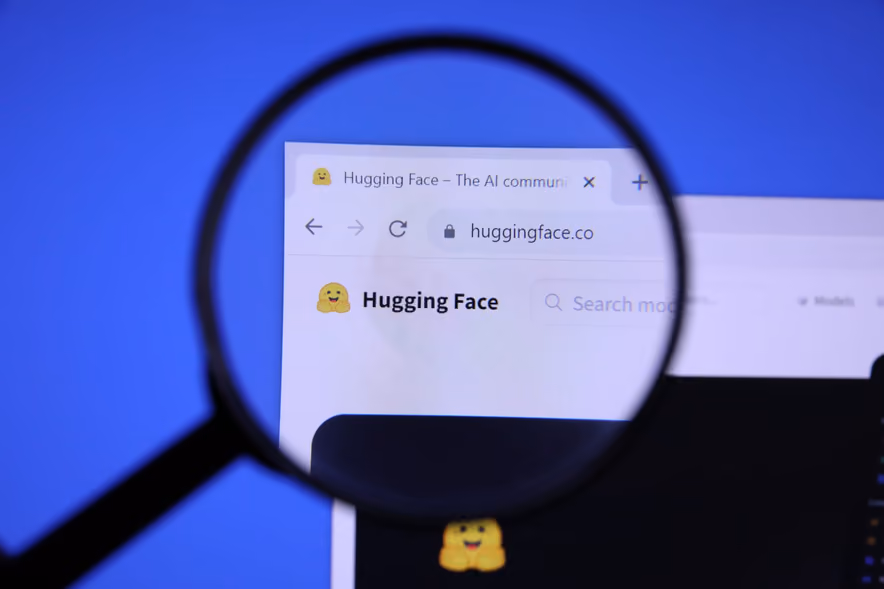 Une loupe posée devant un écran grossit l’en-tête du site huggingface.co, le logo à l’émoji jaune qui sourit bras ouverts à côté du nom Hugging Face, sur un fond bleu.