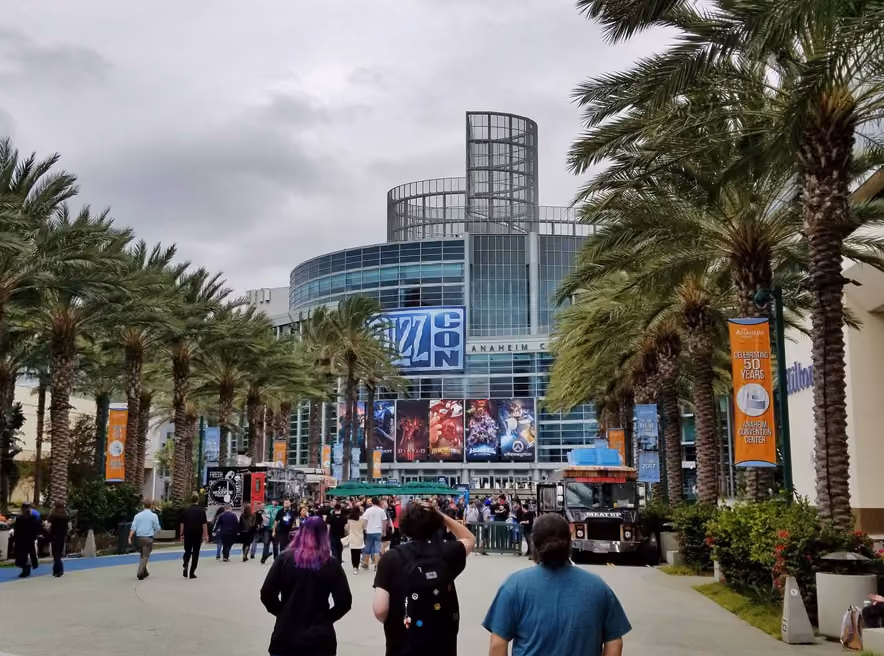 The glass facade of the Anaheim Convention Center on a BlizzCon day, the show's blue logo above posters for Warcraft, Diablo, Hearthstone, and Overwatch, palm trees on both sides, and visitors in black T-shirts walking up the promenade.