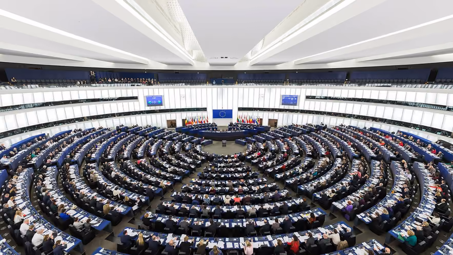 Overhead view of the European Parliament hemicycle in Strasbourg during a plenary session, members seated in concentric rings around a podium below the EU flag and the flags of the member states.