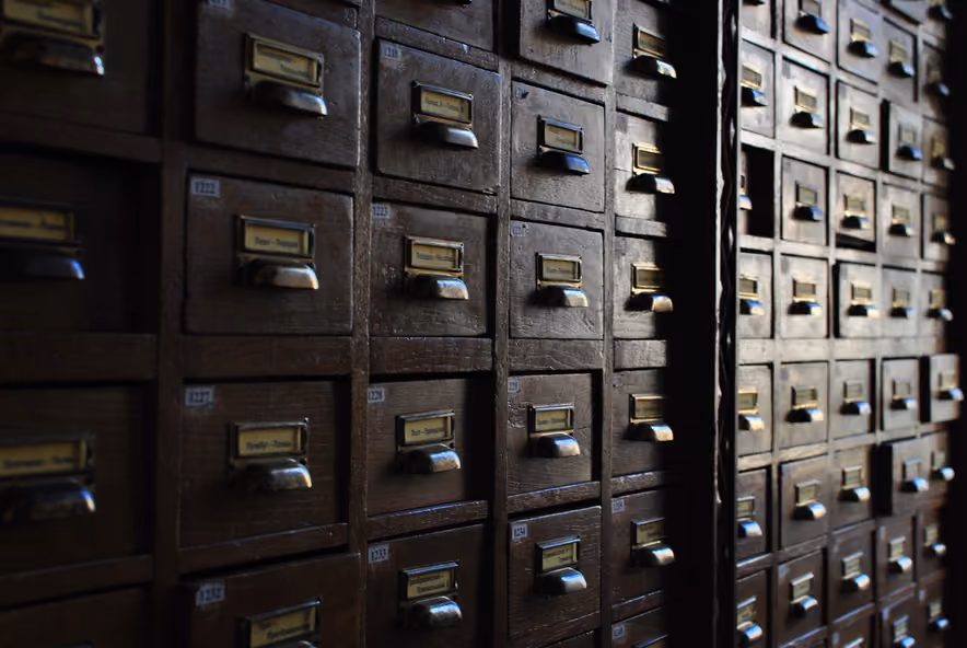 A wall of wooden drawers from an old library card catalog, each with a paper label and a brass pull, receding into shadow on the right.