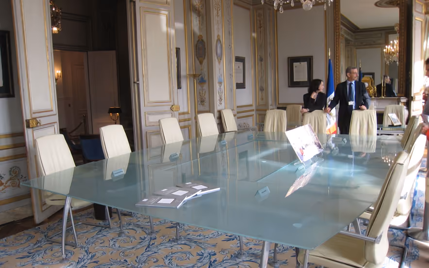 Salle de réunion du Conseil constitutionnel, une longue table de verre dépoli entourée de fauteuils de cuir crème, sous un lustre de cristal, entre des boiseries blanches et or et un tapis à arabesques bleues.