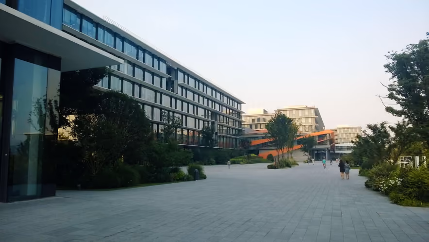 A wide paved plaza on Alibaba's Hangzhou campus, lined on the left by a long glass office building with horizontal awnings, half screened by trees. More glass buildings and an orange walkway close off the far end, where a few figures walk under a pale, hazy sky.