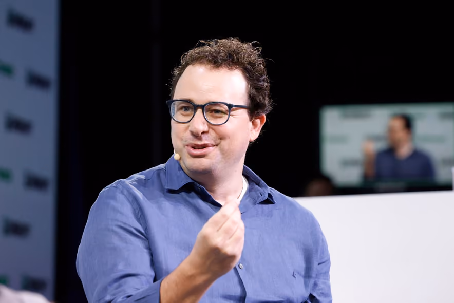 Dario Amodei, in a dark shirt, seated on a stage against a blue backdrop, speaking with his hands spread.