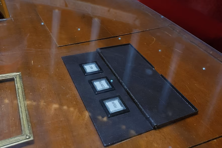 A voting console set into a mahogany desk in the French National Assembly chamber, its three buttons labeled "abst.", "contre" and "pour", in front of a red velvet bench.