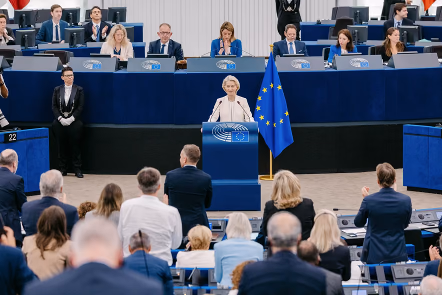 Ursula von der Leyen, in a white suit, standing at the podium of the European Parliament chamber beside the blue star-spangled flag, in front of the presiding bench, while members in the foreground give her a standing ovation.