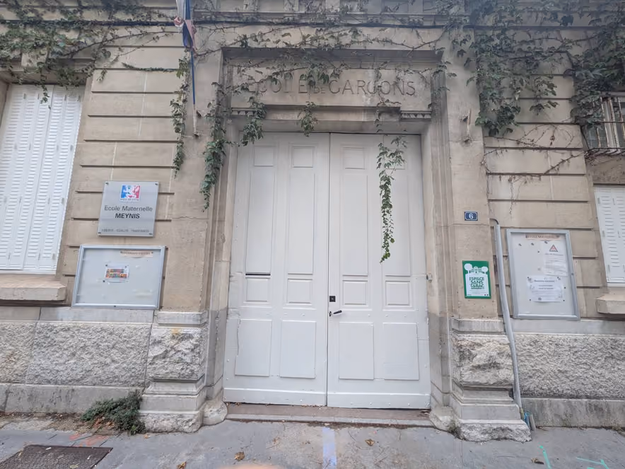 Façade en pierre d’une ancienne école de garçons devenue école maternelle, avec une grande porte blanche à deux battants, du lierre qui retombe du linteau, une plaque « École maternelle Meynis » et deux panneaux d’affichage vitrés garnis de feuilles de papier.