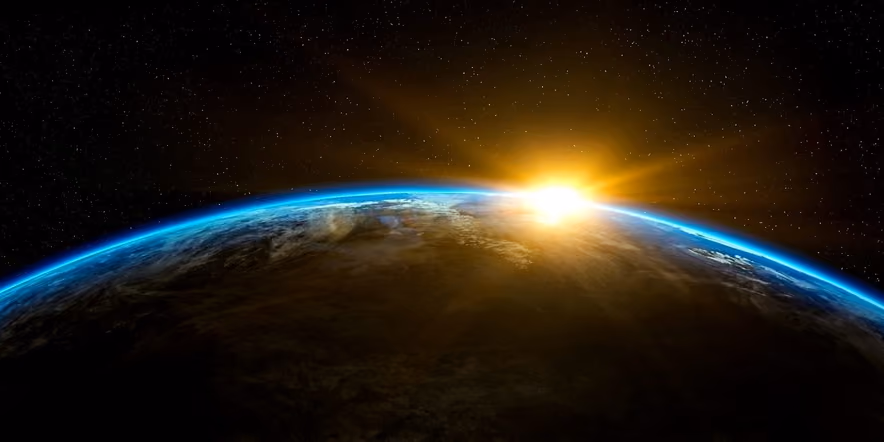 The curve of the Earth seen from space, a bright sunrise on the horizon, the atmosphere traced by a thin blue rim against a starry sky.
