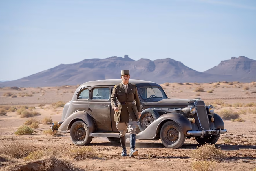 Simon Abkarian in a general's uniform and kepi, walking past a black 1940s car parked in the middle of a desert, barren mountains on the horizon.