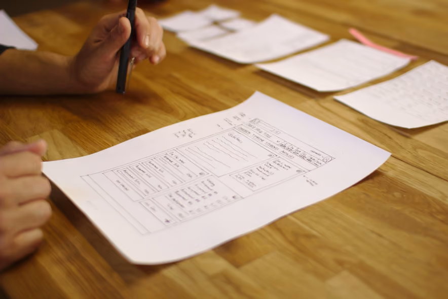 A sheet of paper on a wooden table with a website screen drawn on it in pen, tabs, columns, and lists, under the hands of its designer, who holds a marker.