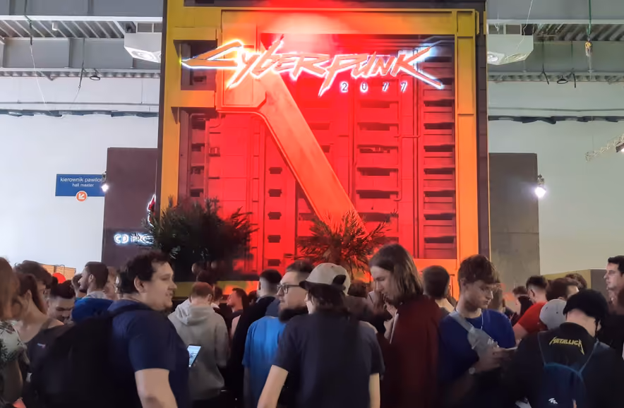 A dense crowd of convention visitors, nearly all seen from behind, waits in front of a huge booth topped by the red and white neon sign of Cyberpunk 2077, mounted on a fake apartment facade.