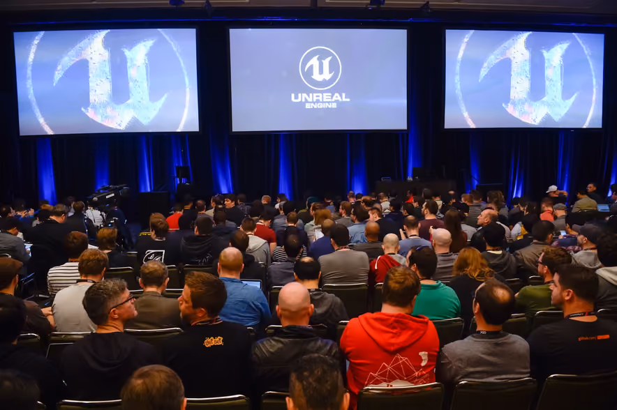 Seen from behind, a packed room of several hundred people seated before three large screens under blue curtains, the middle one showing the Unreal Engine logo and the other two its giant U.