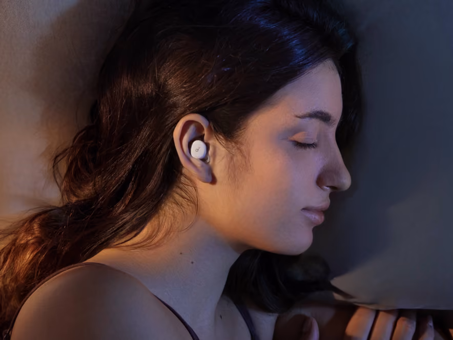 A pair of white earbuds built for sleeping, lying flat beside their oval charging case on a light background.