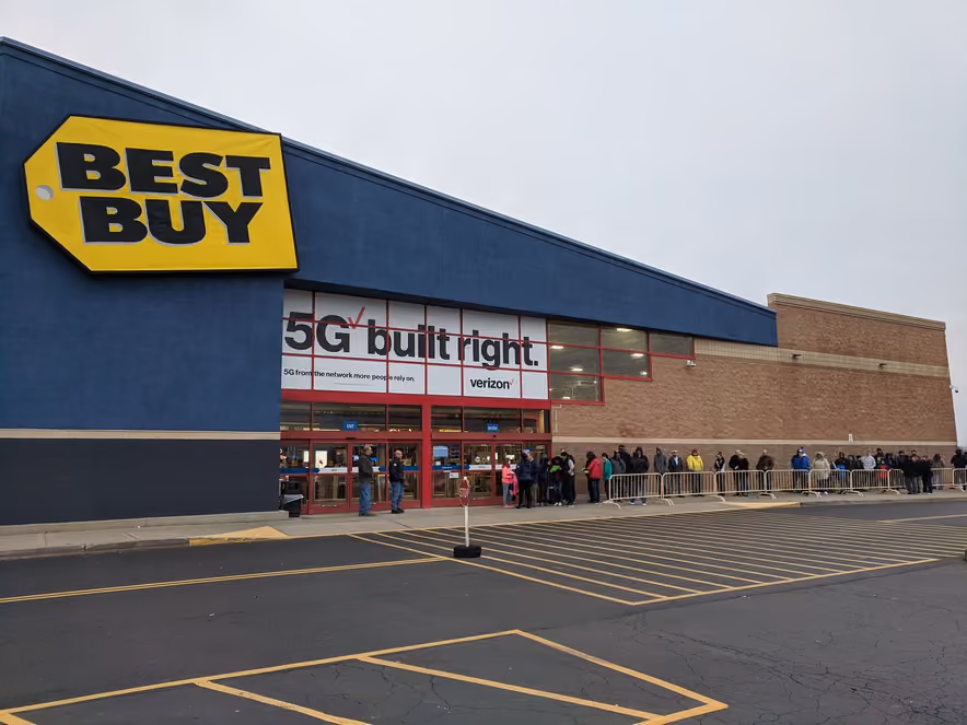 A long line of bundled-up shoppers waits under a gray sky outside a Best Buy entrance, below its yellow sign, beside a mostly empty parking lot.