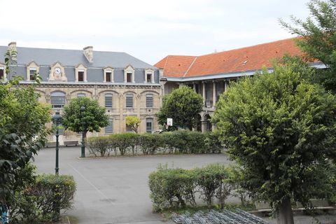 An empty high school courtyard, a wide paved yard edged with hedges and shrubs, facing a pale stone building topped by a clock and a long wing under orange tiles, with a basketball hoop and empty bike racks.