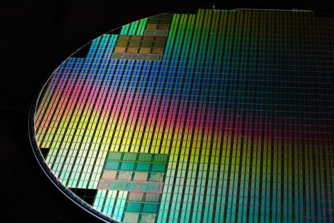 Close-up of a tilted silicon wafer covered in hundreds of rectangular chips, throwing iridescent green, pink, and blue reflections against a black background.