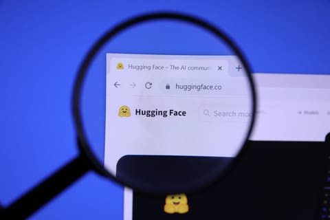A magnifying glass held in front of a screen enlarges the header of huggingface.co, the yellow open-armed smiling emoji logo next to the Hugging Face name, against a blue background.