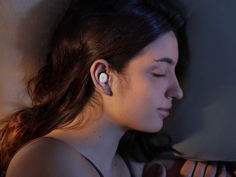 Une paire d’écouteurs blancs conçus pour le sommeil, posés à plat à côté de leur boîtier de charge ovale sur fond clair.