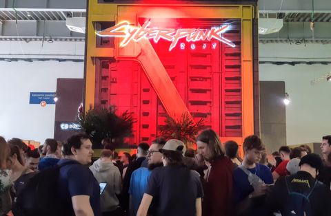 Une foule compacte de visiteurs, presque tous de dos, patiente devant un immense stand de salon dominé par l’enseigne lumineuse rouge et blanche de Cyberpunk 2077, montée sur une façade d’immeuble factice.