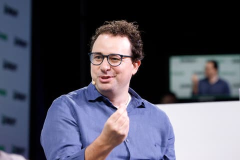 Dario Amodei, en chemise sombre, assis sur une scène devant un fond bleu, parle en écartant les mains.