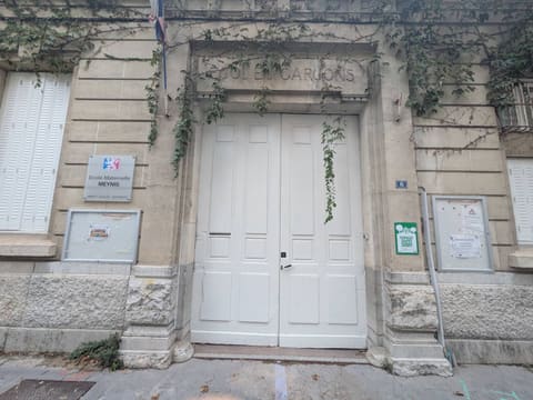 Façade en pierre d’une ancienne école de garçons devenue école maternelle, avec une grande porte blanche à deux battants, du lierre qui retombe du linteau, une plaque « École maternelle Meynis » et deux panneaux d’affichage vitrés garnis de feuilles de papier.