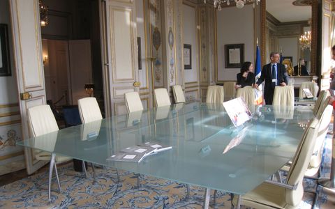 Salle de réunion du Conseil constitutionnel, une longue table de verre dépoli entourée de fauteuils de cuir crème, sous un lustre de cristal, entre des boiseries blanches et or et un tapis à arabesques bleues.