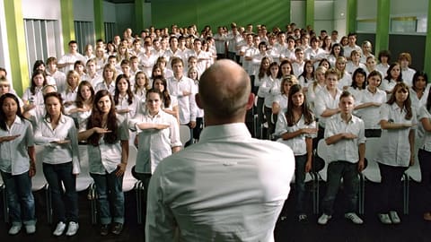 Face à un professeur de dos en chemise blanche, une salle entière d'élèves en chemise blanche exécute le même salut, bras droit tendu à l'horizontale sur la poitrine.
