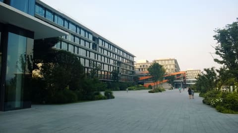 Vaste esplanade pavée du campus d’Alibaba à Hangzhou, bordée à gauche par un long immeuble de bureaux en verre aux auvents horizontaux, à demi masqué par des arbres. D’autres bâtiments vitrés et une passerelle orange ferment le fond, où quelques silhouettes marchent sous un ciel blanc et voilé.